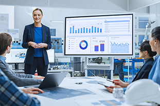 Modern Industrial Factory Meeting: Confident Female Engineer Uses Interactive Whiteboard, Makes Report To A Group Of Engineers, Managers Talks And Shows Statistics, Growth And Analysis Information
